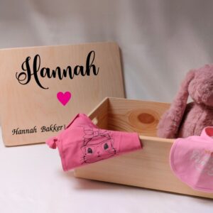 Houten opbergdoos met naam en geboortegegevens, voorbeeld met naam 'Hannah', roze hart, babykleding en knuffel in en naast de bewaardoos.